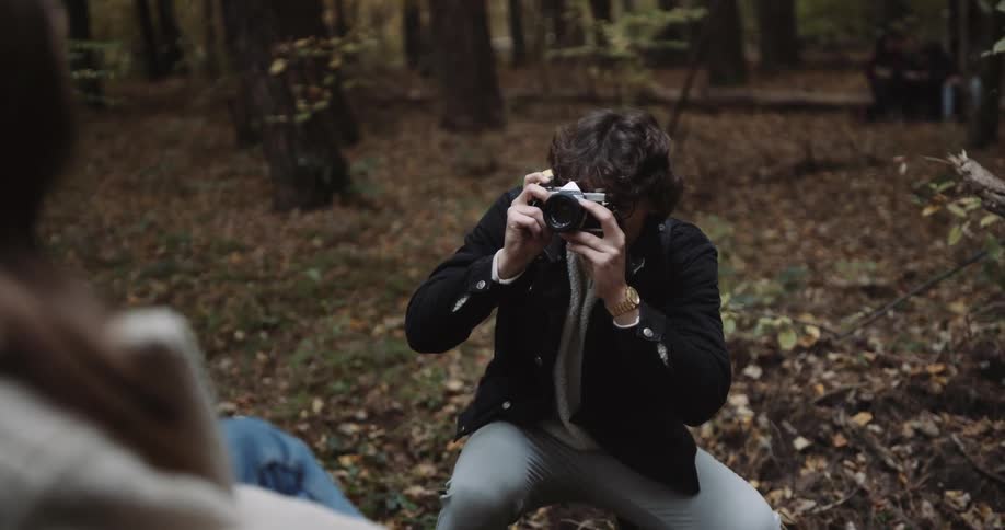A young woman is sitting and posing for a man taking photos in a woodland area during the day in the fall season