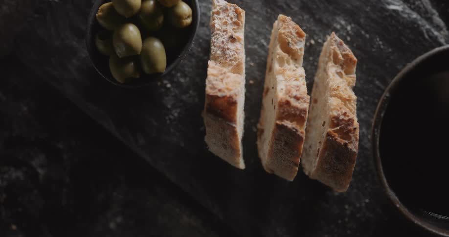 Bread slices sit on board between bowl full of liquid and bowl containing olives