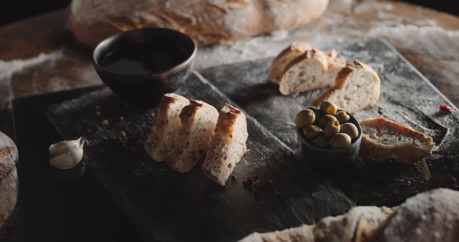 Loafof bread sits on table near board holding bread slices olives and a full bowl