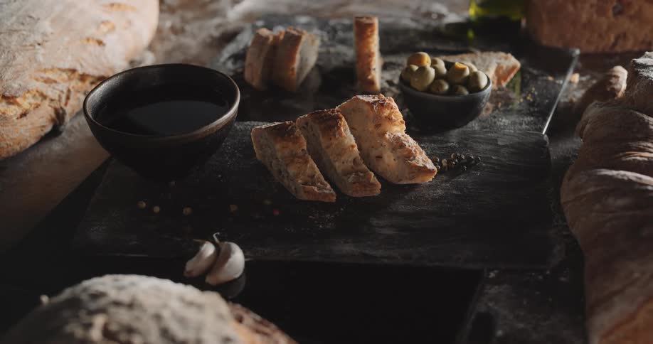 An assortment of bread whole and slices next to a bowl of olives and cloves of garlic