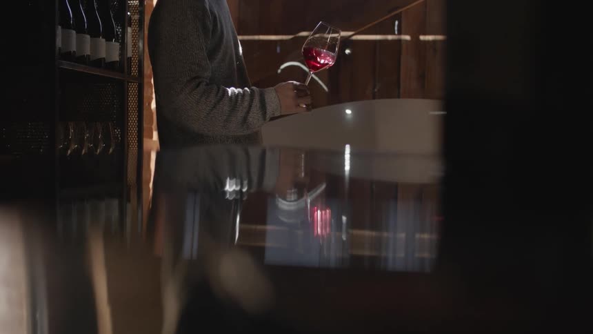 A man is standing indoors near a wine collection while holding a wine glass and swirling the red wine around in the glass in the daytime