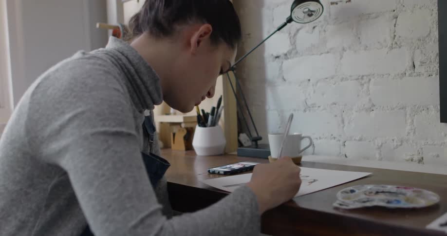 A young female artist is sitting indoors and painting a picture