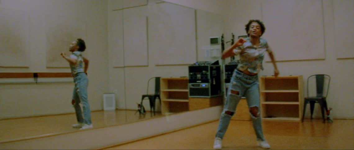 Female dancer wearing blue shirt and ripped jeans dances in front of large mirrors in room