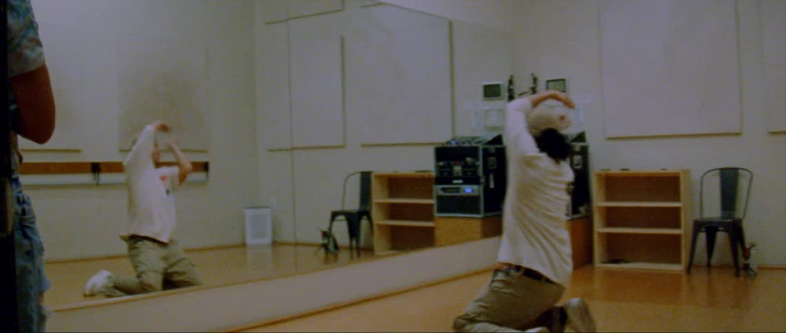 Man wearing brown hat white t shirt and pants dances in front of large mirrors in room