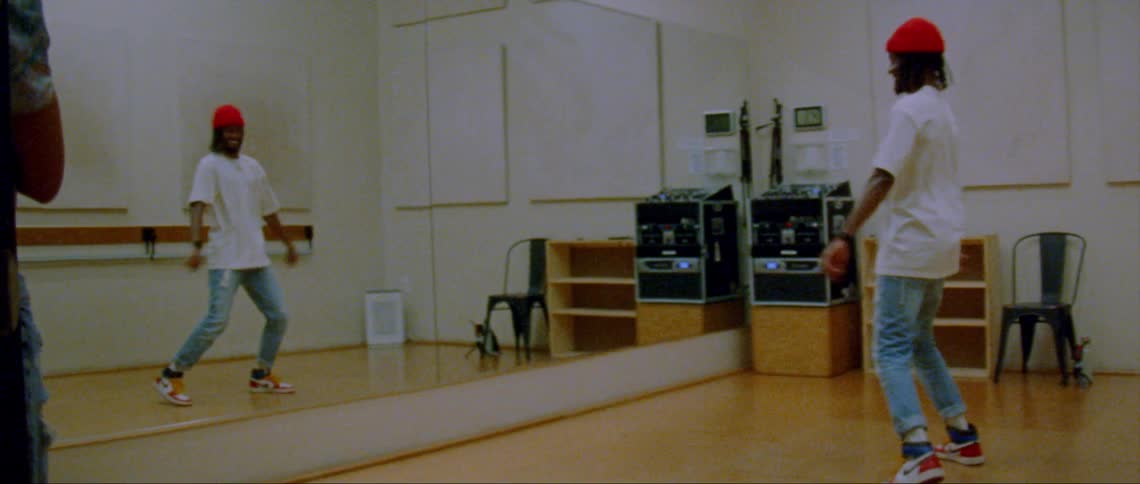 A man wearing a white shirt and orange cap dancing in front of a mirror