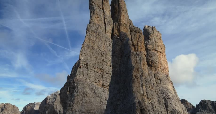 A person is climbing up the side of a huge rock formation in the badlands under a cloudy sky marked with contrails