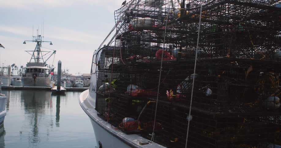 Boat with stacked metal cages bobs at dock across from marina under cloudy sky