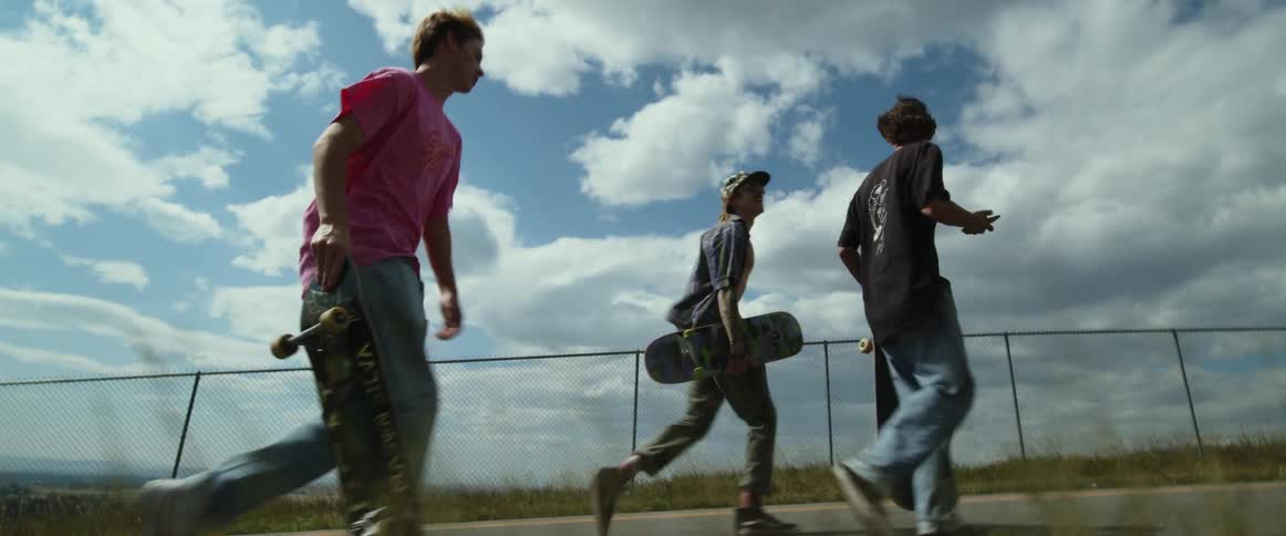 Three teenage boys are carrying their skateboards as they walk uphill on a road and one boy is looking at his cell phone