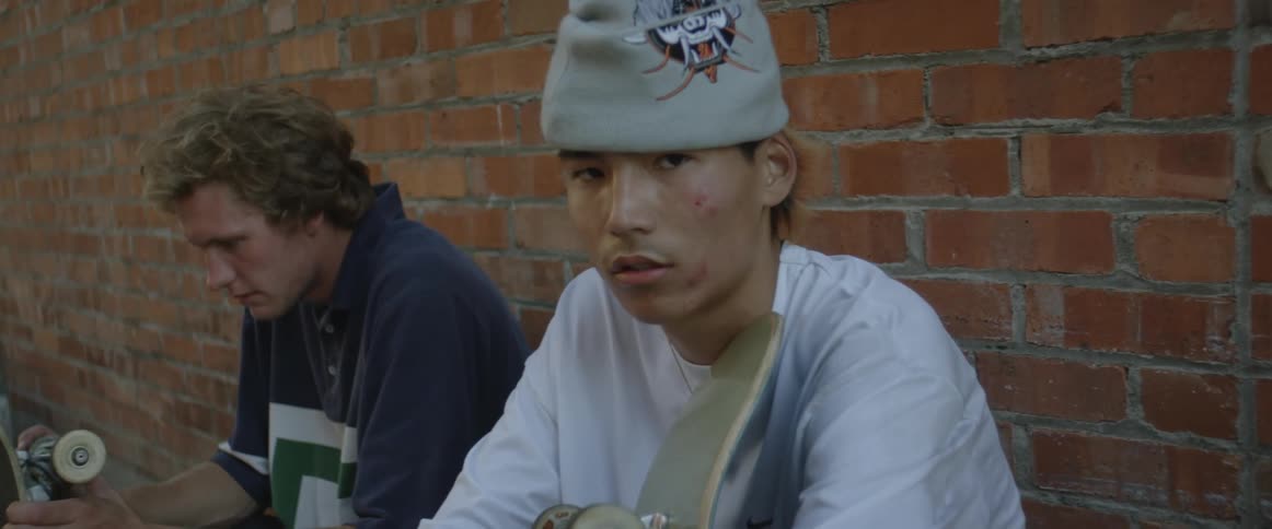 Two teenage boys are sitting near a brick wall as they hold their skateboards