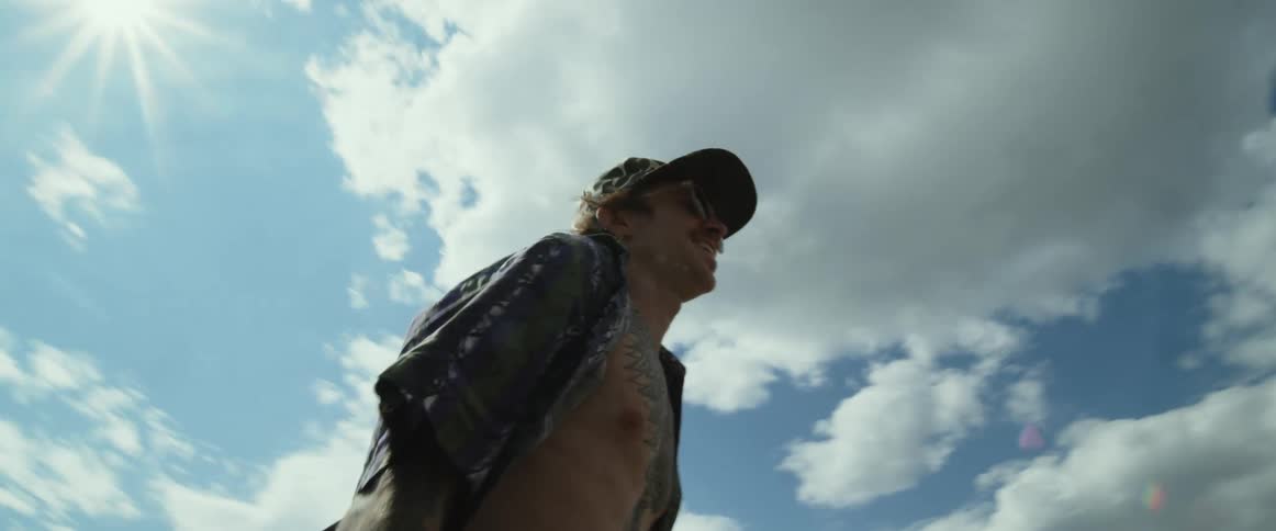 Man wearing cap sunglasses unbuttoned shirt and tattos walks down the street carrying skateboard