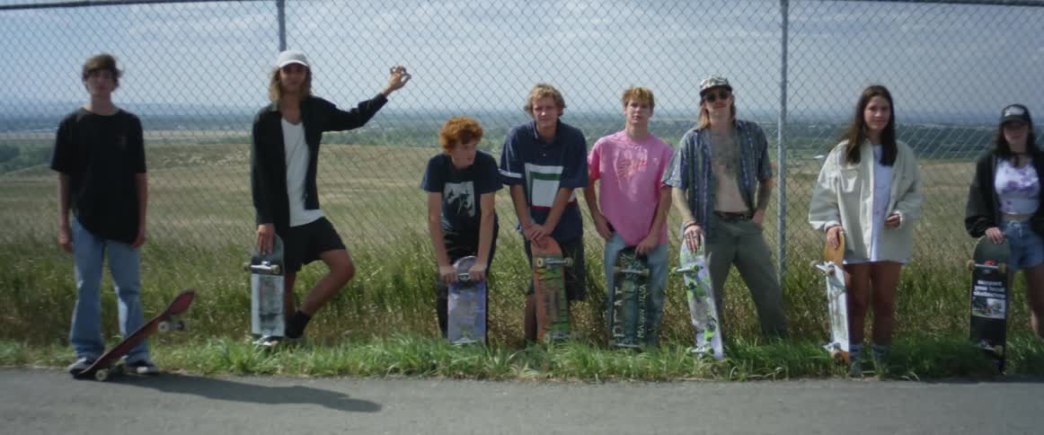 A small group of male and females standing in front of a chain linked fence with skateboards in their hands
