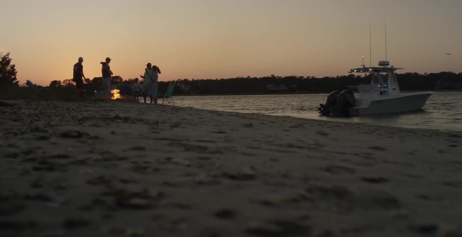 Friends gather around a campfire on the coast next to a boat at evening