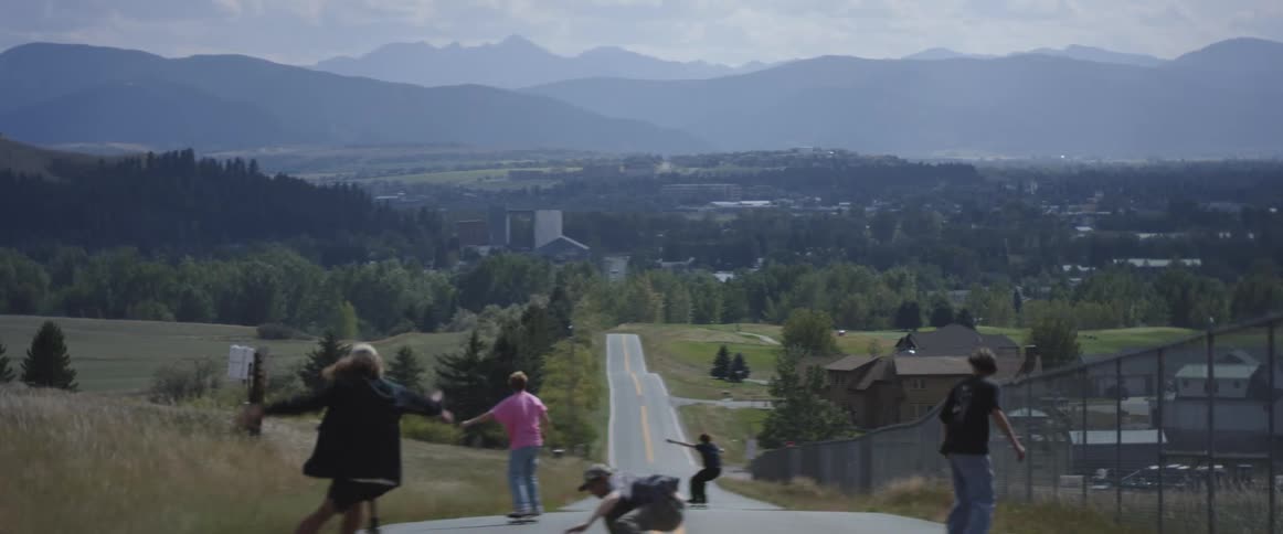 A group of teenagers ride skateboards downhill past a neighborhood