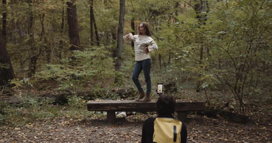 Man takes video with smartphone as woman poses on bench in woods