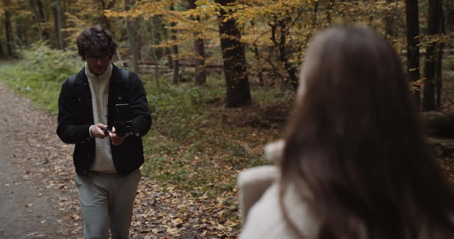 A teenage couple are hiking through the woods and he is holding his cell phone on a stick as he takes her picture