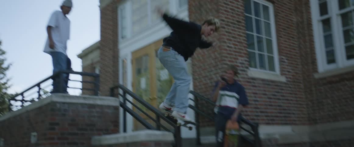 A teenage boy stands on a ledge over a hand rail and rides his skateboard down the rail before losing his footing on the steps
