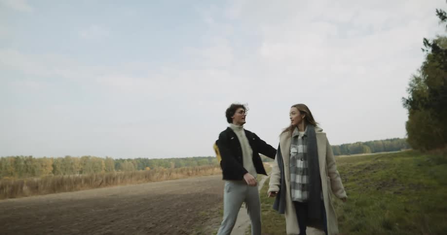 A couple are holding hands while running along a dirt path near farmland and then into a wooded area in the daytime