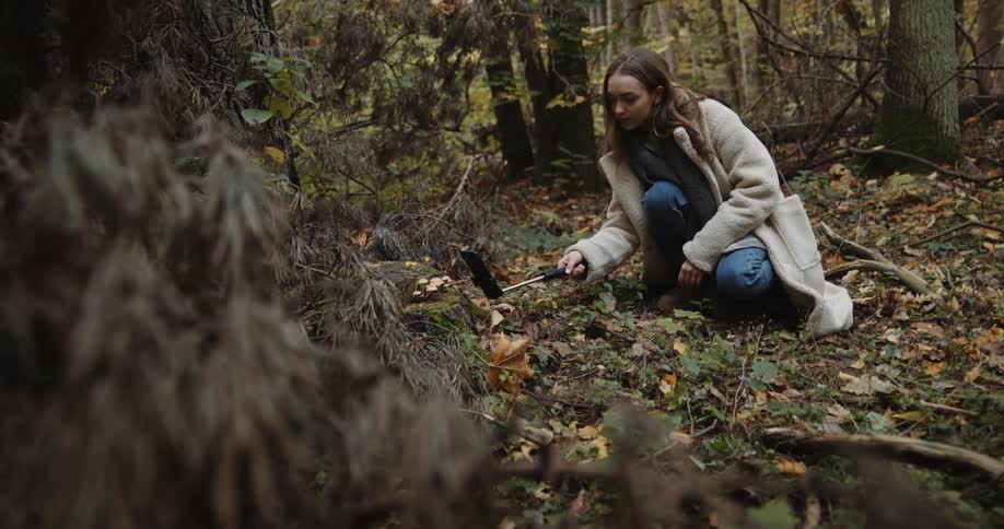 Young woman crouching in the forest searchs around with a small metal detector