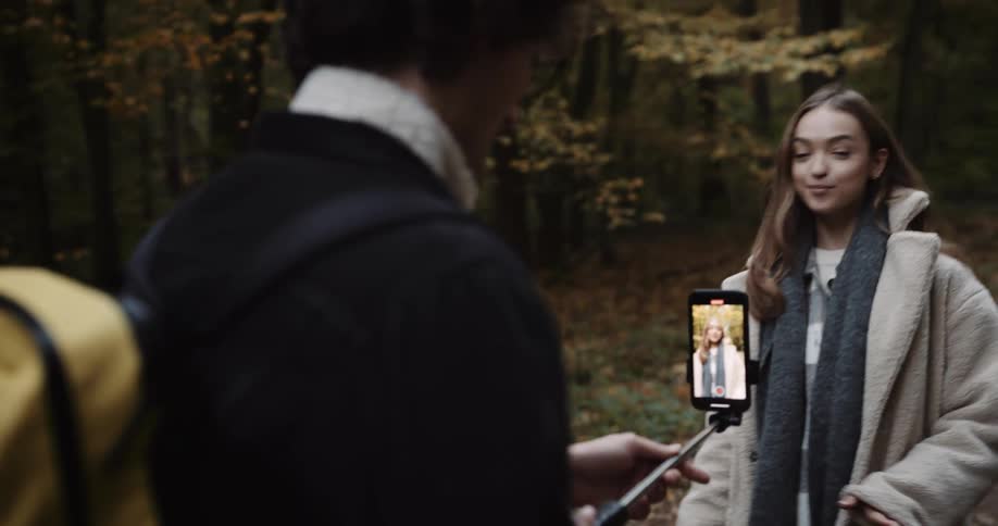 Two teenagers are walking through the woods and he is walking backwards as he holds a cell phone on a selfie stick and records her walking and talking