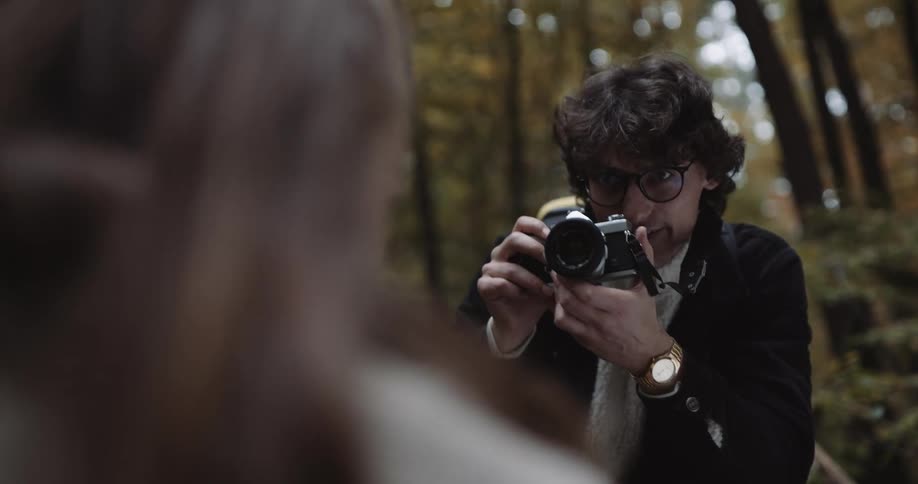 A young woman is seated and posing while a photographer is taking photos in a woodland during the day