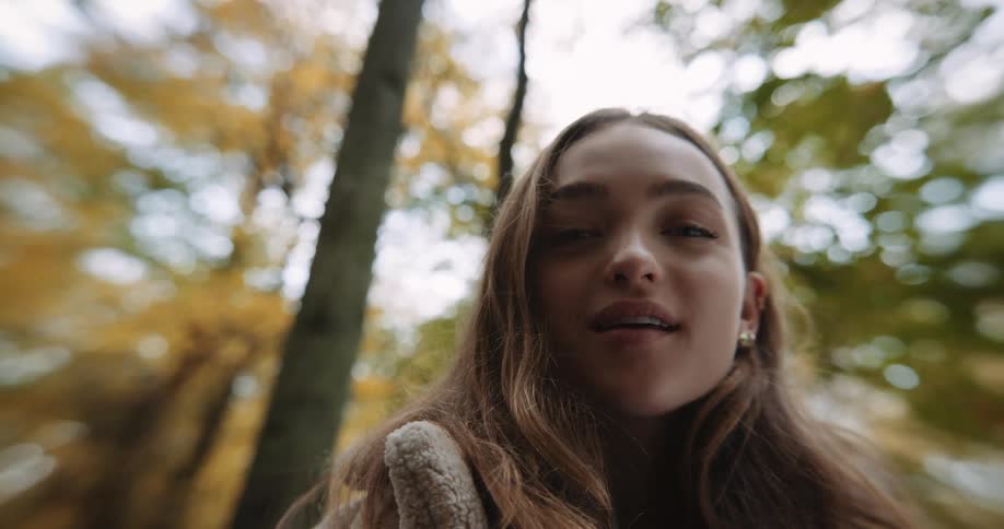 Girl with brown hair and beige coatand earrings looks around and looks down at camera in the forest autumn