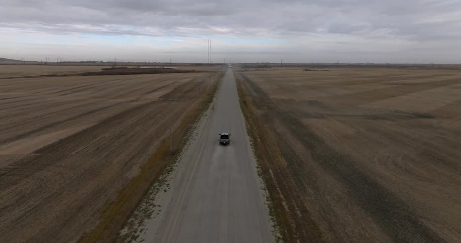 An aerial view of a truck traveling on a long and straight dirt road with flat farmland on both sides on a cloudy day