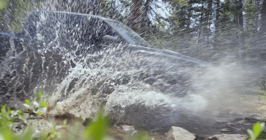 A pickup truck splashes water as it crosses a stream in a forest on a sunny day