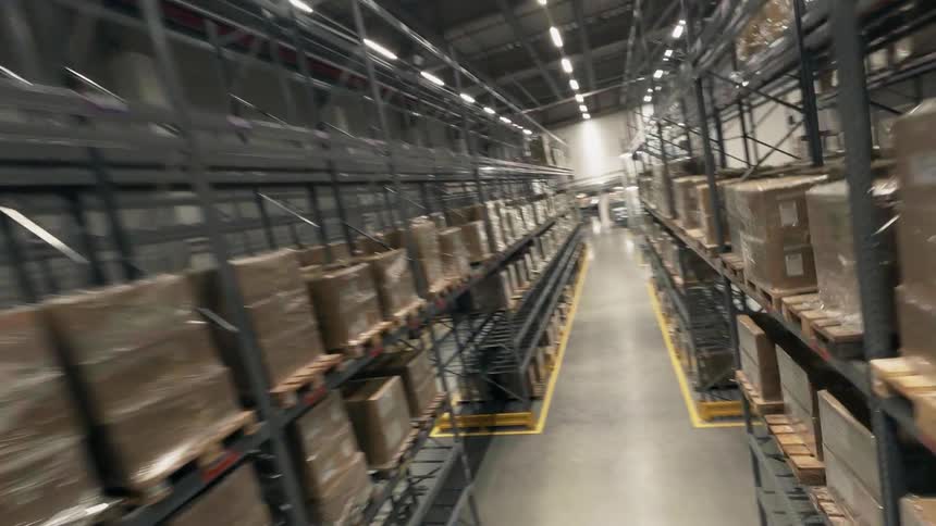 An FPV drone shot floating through the Interior of a large warehouse with metal storage shelves holding boxes of shipments and goods