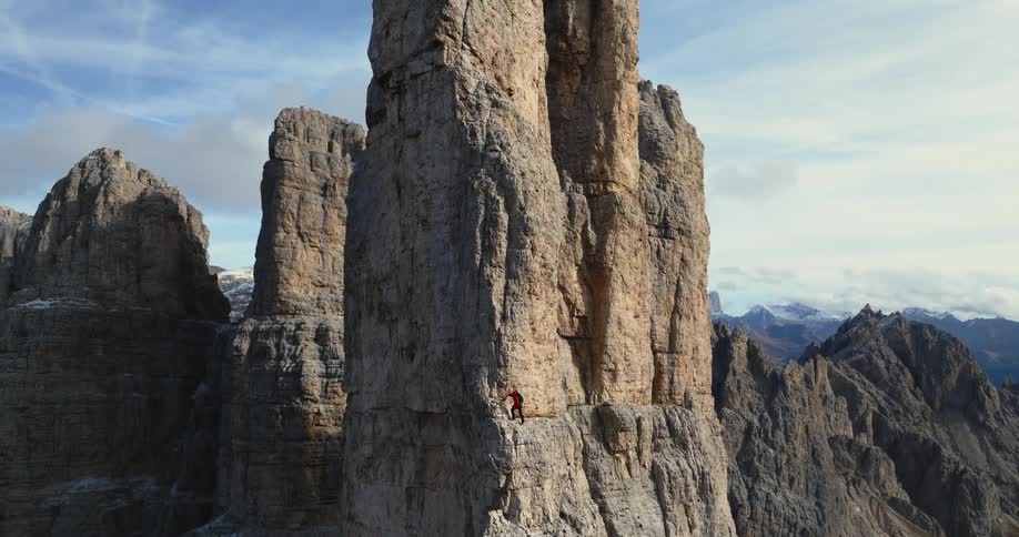 Person climbs sheer side of rock formation in mountain range under a cloudy sky