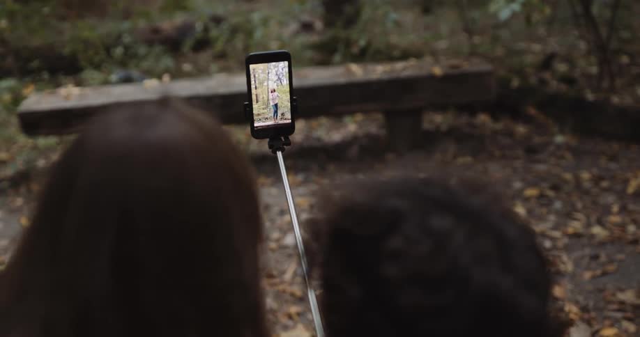 Two teenagers are standing in the woods holding their phone on a selfie stick as they watch a video they made