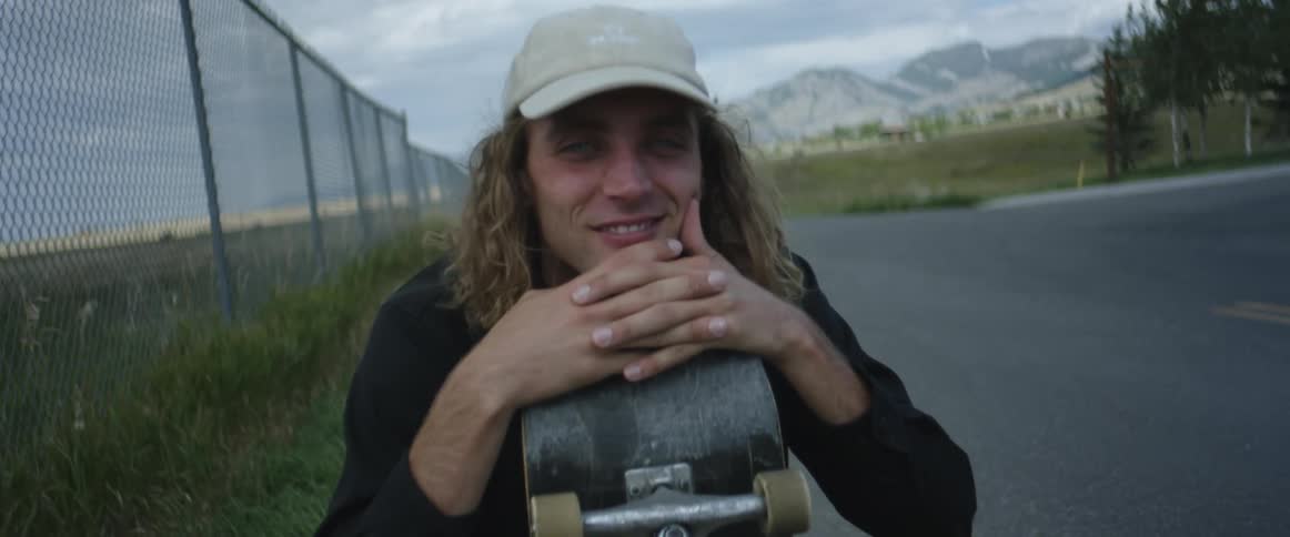 Young long haired man holds a skateboard while sitting a the side of a road