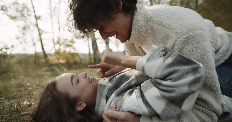 A teenage couple are lying on the ground near the woods talking and laughing as he touches her nose with his finger