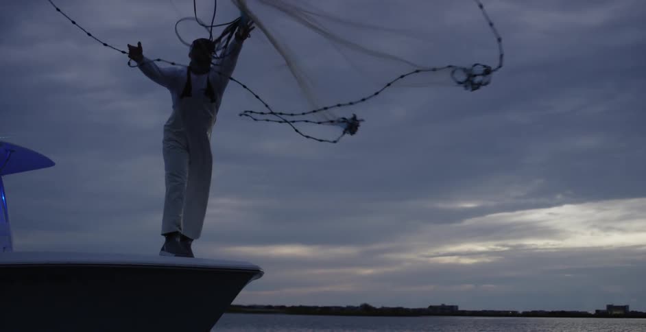A man standing on a boat is casting a large fishing net on a lake in the early morning of a cloudy day