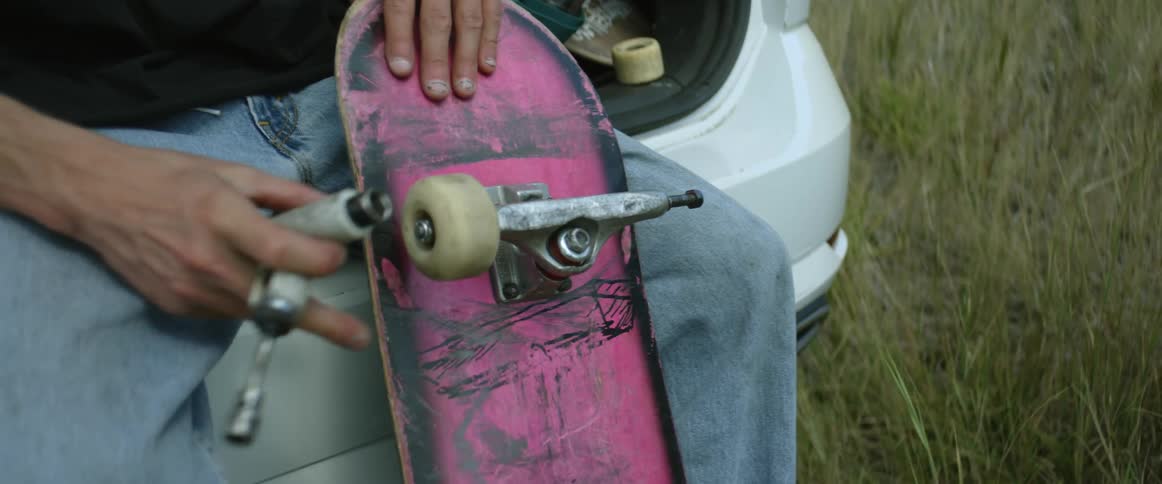 A teenage boy is sitting on the back of a car holding a wrench as he fixes the wheels on his skateboard