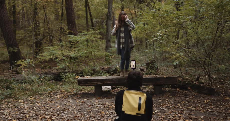 Woman walks to bench in woods and steps onto it then poses as man takes pictures with a smartphone