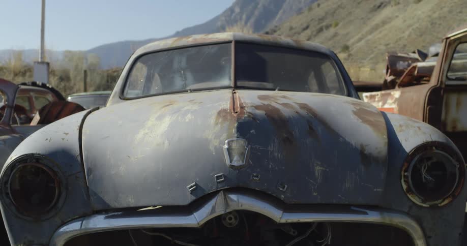 The front fender and windshield of an old blue car in a junk yard with mountains in the background