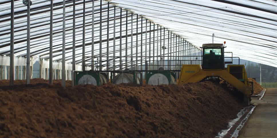 Operator sits in large commercial machinery as it tills long mound of dirt in sheet roofed structure