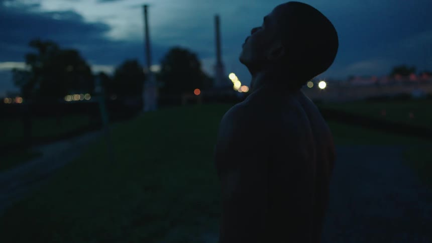 An athletic man stands shirtless with his eyes closed near a public sports field just after sunset