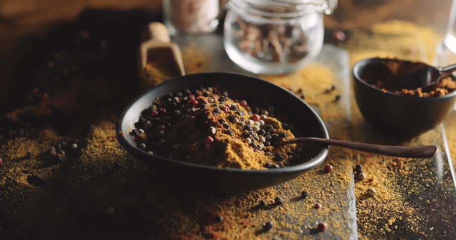 Bowl of spices and peppercorns rests board and surface both scattered with spilled spices and peppercorns