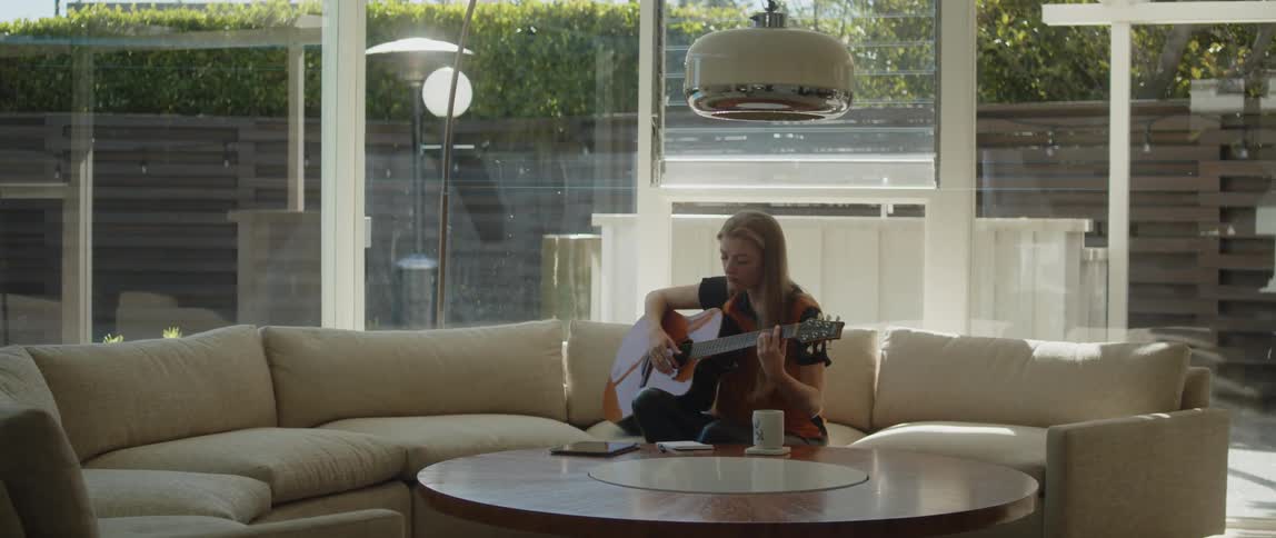 A woman idly strums a guitar while sitting on a sofa in the living room of a house during the day