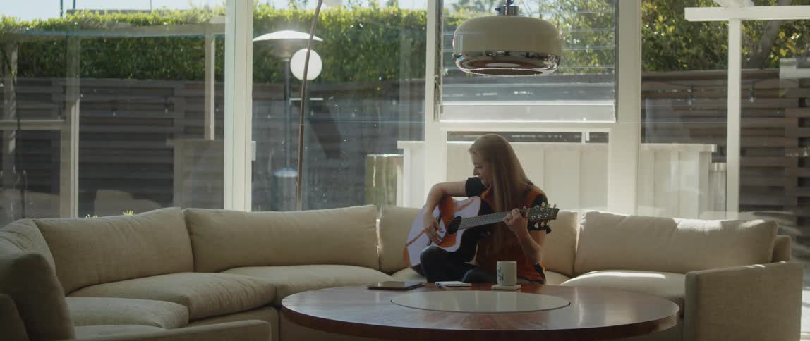 A woman writes on a notepad before beginning to strum a guitar while sitting on a sofa in the living room of a house during the day