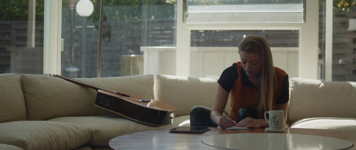 Woman sits on couch near a guitar writing on a notepad