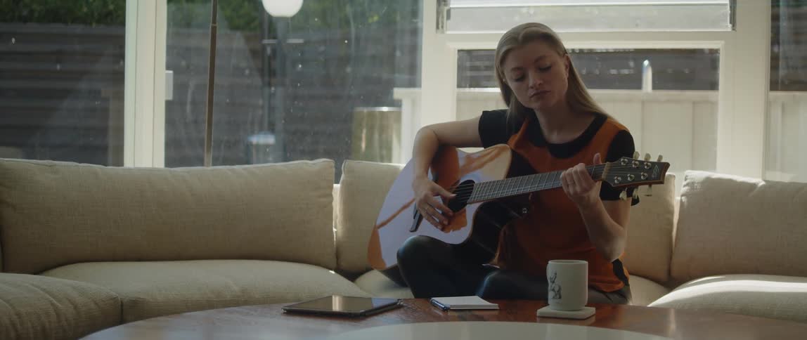 Woman with brown hair black and orange shirt plays acoustic guitar sitting on beige couch in the living room
