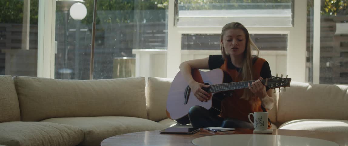 A female sitting on the couch singing and strumming a guitar