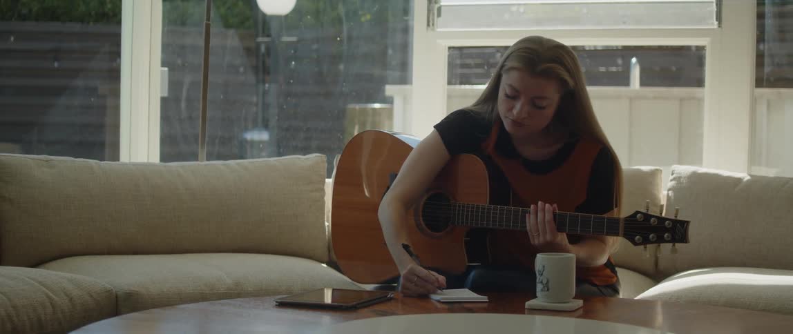 A woman is sitting on the living room couch playing an acoustic guitar when she stops to write something on paper sitting on the table in front of her then continues to play