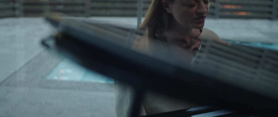 Blonde woman wearing necklace white sweater plays grand piano next to window overlooking pool and fences