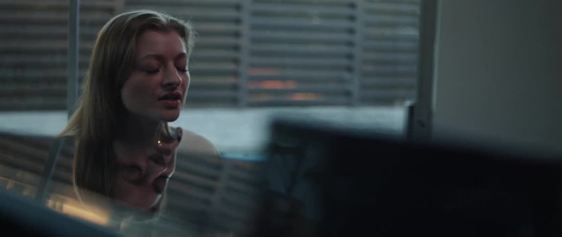 Blonde woman wearing white sweater plays grand piano and sing a song next to windows with blinds in room at home