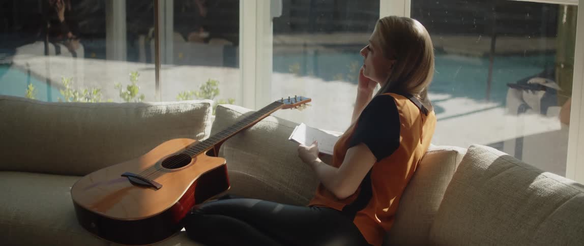 Blonde woman in black and orange shirt and black pants sits on beige couch thinking and writing in notebook in living room at home