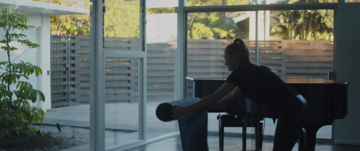 A piano in the corner while a woman walks over and places a yoga mat on the floor