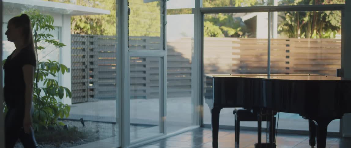 A woman is standing in a room of windows near a piano as she looks outside and walks away